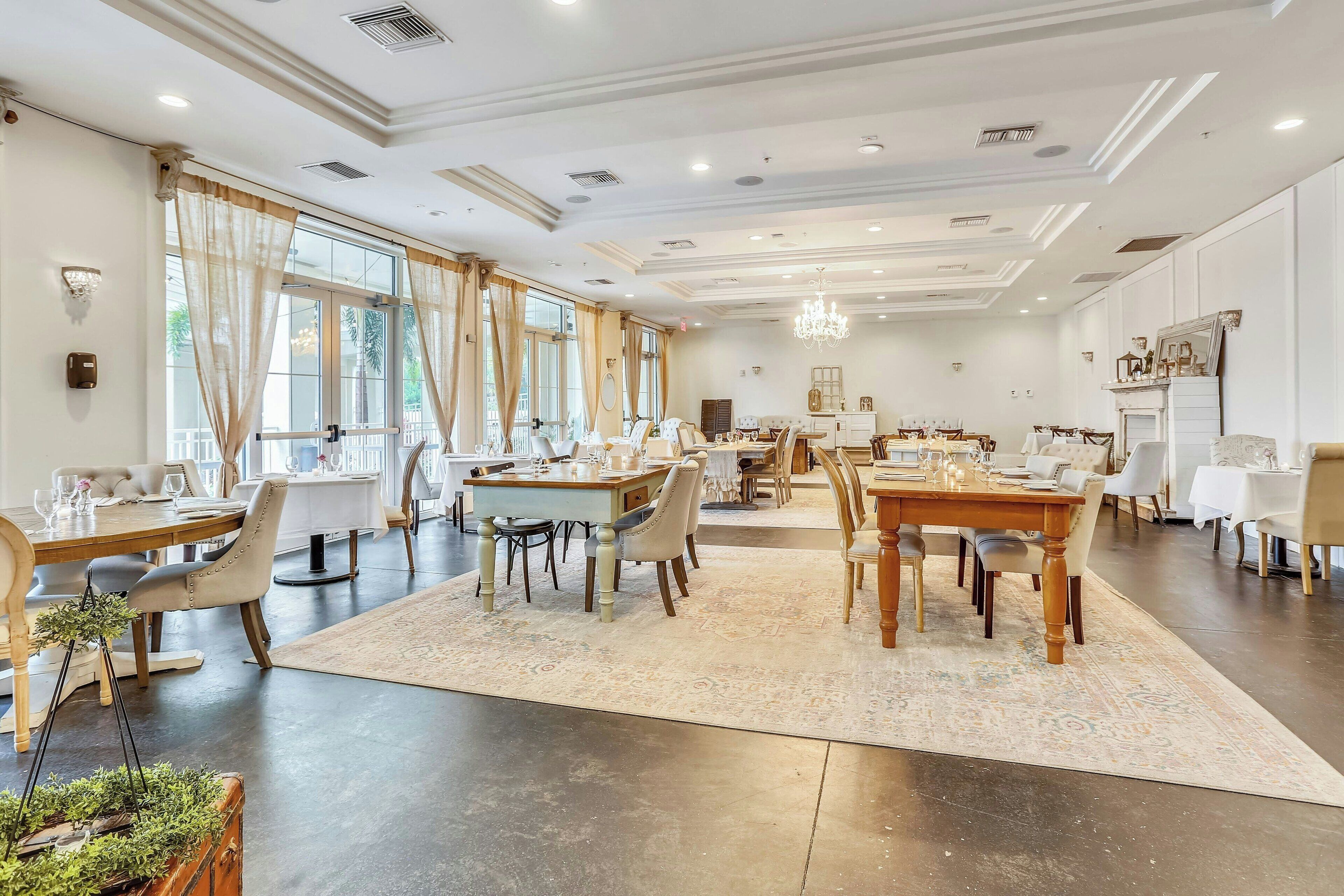 Un comedor amplio con varias mesas y sillas, ventanas grandes con cortinas y un candelabro.