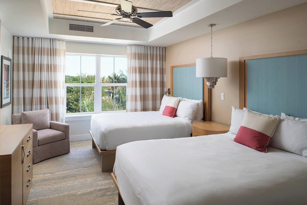 Hotelkamer met twee bedden, een stoel, een dressoir, een plafondventilator en een raam met gordijnen.