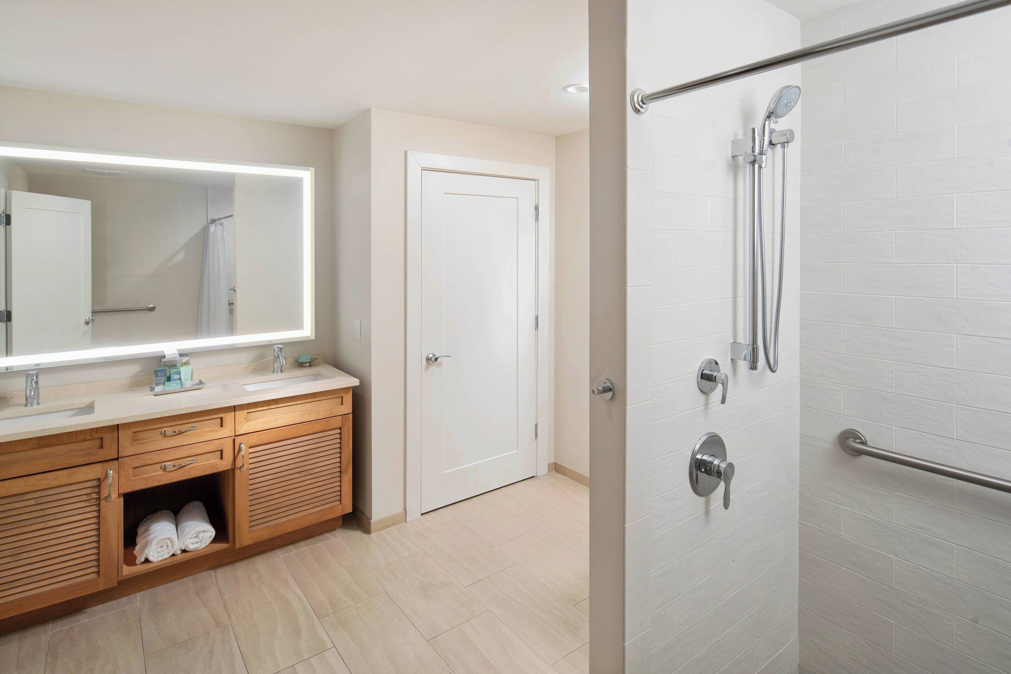 A modern bathroom with a large mirror, wooden vanity, two sinks, a shower with a glass door, and a towel rack.