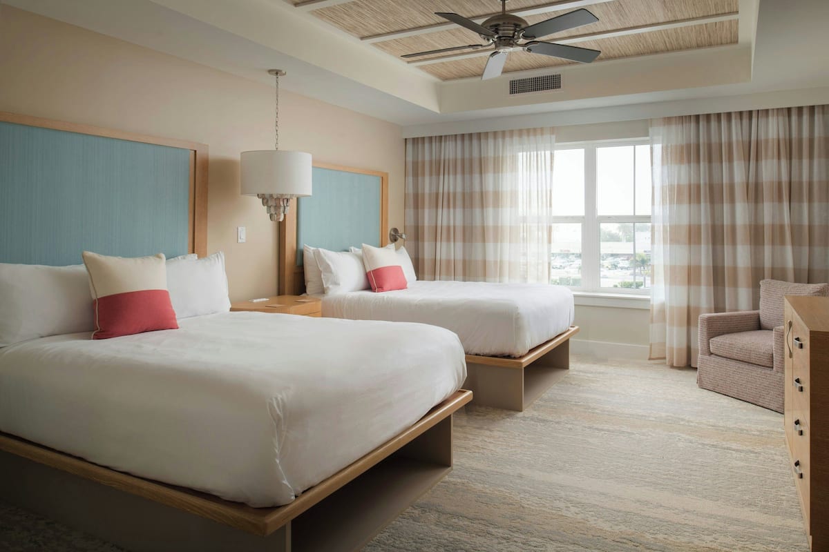 Een hotelkamer met twee bedden, een plafondventilator, een raam met gordijnen en uitzicht op het omliggende landschap.