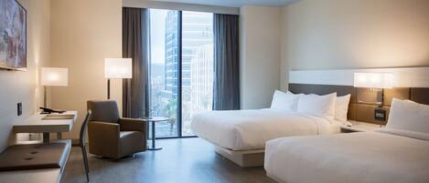 A hotel room with two beds, a desk, a chair, and a view of the city through large windows.