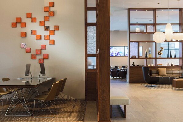 Un moderno vestíbulo de hotel con una mesa de conferencia de vidrio, detalles de madera y una zona de estar con un sofá y sillones.