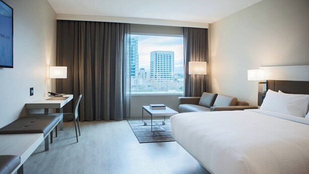 A hotel room with a large bed, a desk, a chair, a small table, and a view of the city.