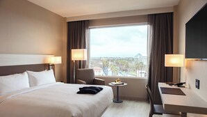 Ein Hotelzimmer mit einem großen Bett, zwei Nachttischen inklusive Lampen, einem Schreibtisch mit Stuhl und Blick auf die Stadt durch große Fenster.