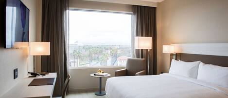 Habitación de hotel con una cama grande, dos sillas, un escritorio y una mesita. Se observa una ventana con cortinas, un televisor montado en la pared y dos lámparas a ambos lados de la cama.