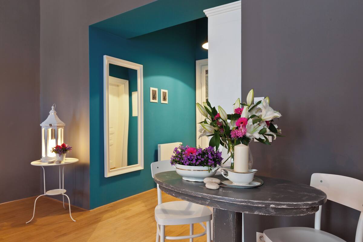 Een eetgedeelte met een muur in teal, een witte stoel, een ronde tafel met een bloevas, en een klein wit tafeltje met een lantaarn.