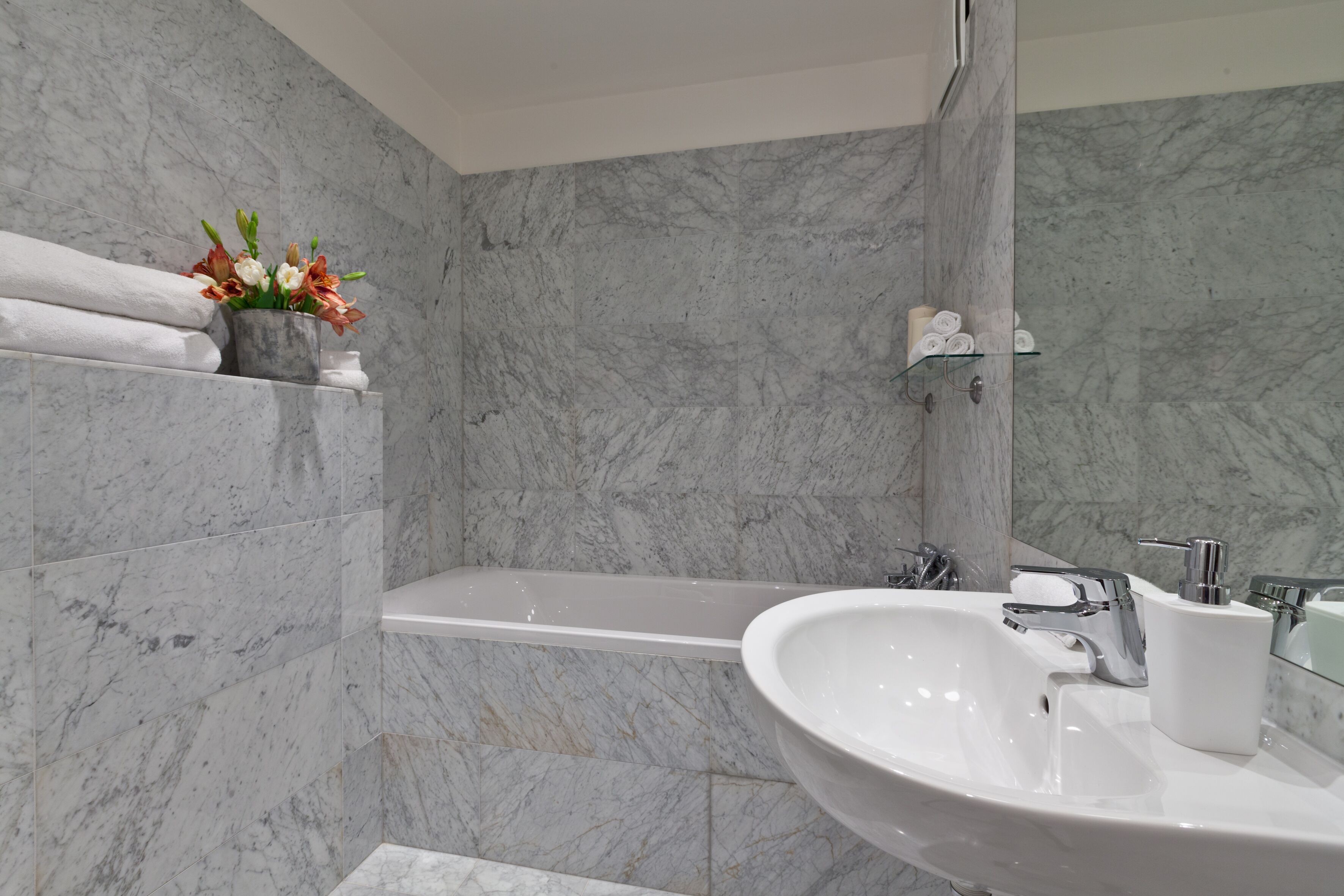Un baño con pared de mármol, lavabo blanco, bañera y estante con toallas y flores.