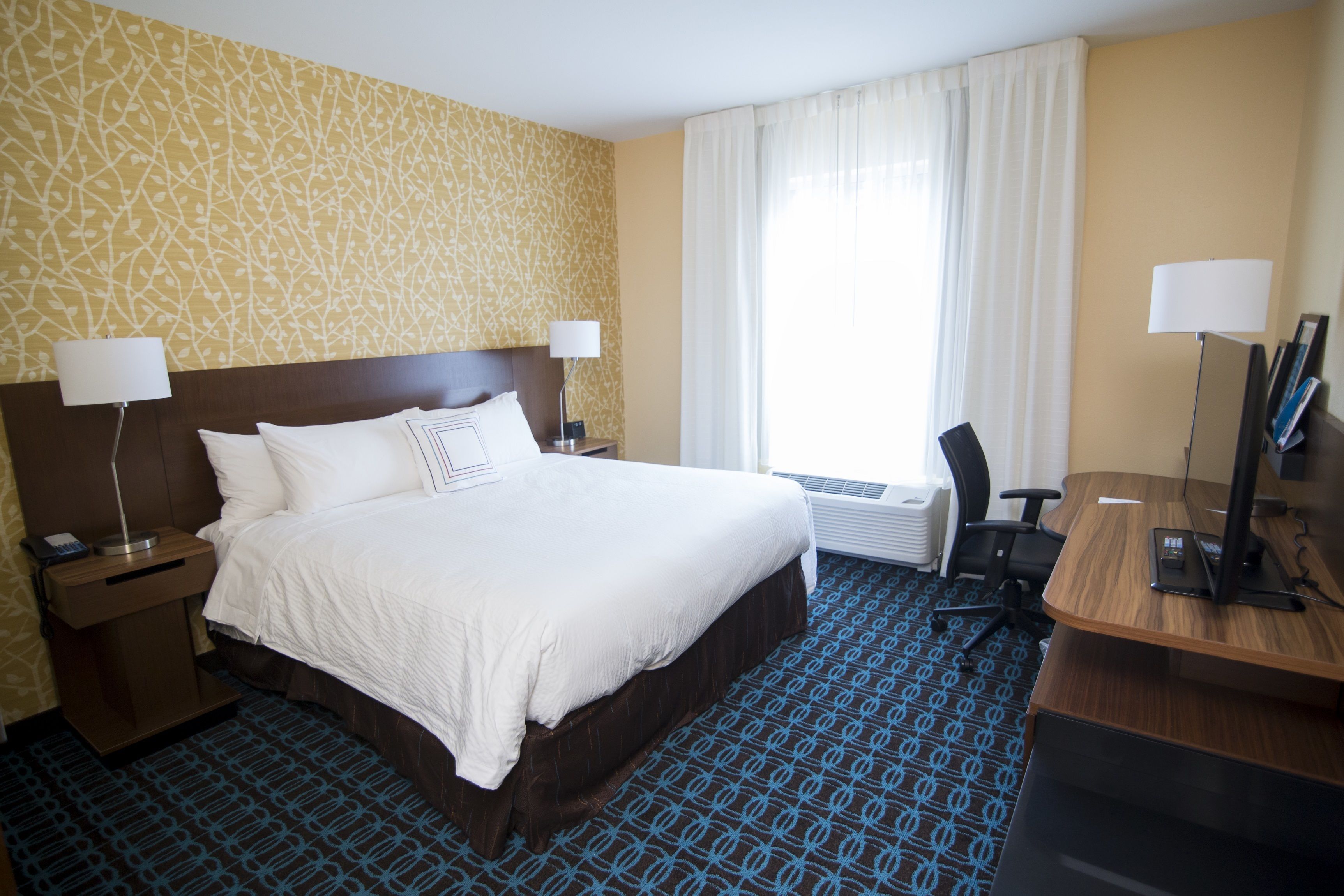Quarto de hotel com uma cama grande, duas mesas de cabeceira com luminárias, uma escrivaninha com computador e uma janela com cortinas.
