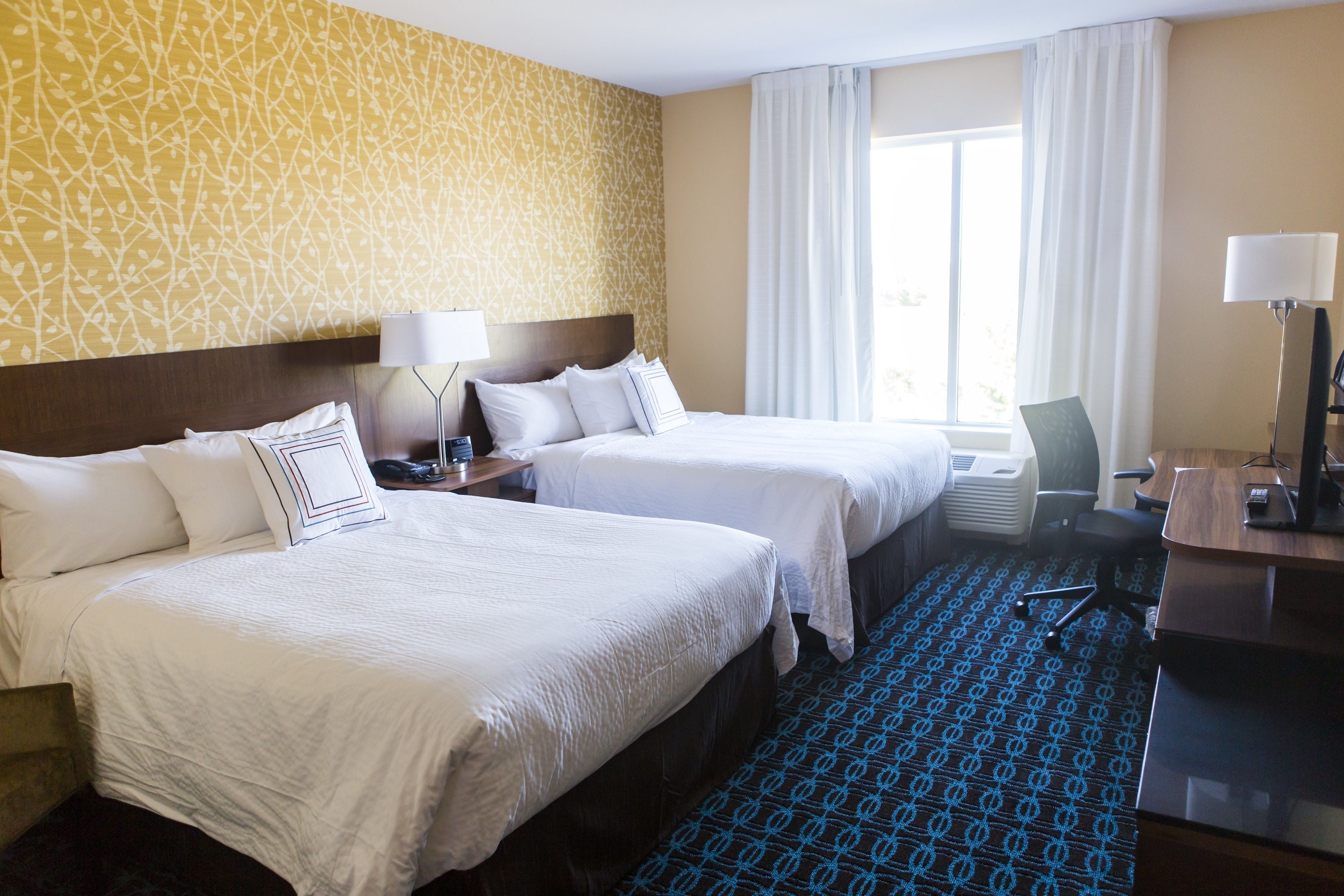 A hotel room with two beds, a desk, a chair, a lamp, and a window with curtains.