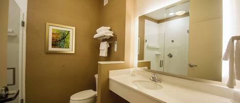 Un baño moderno con pared color beige, inodoro blanco, lavamanos con un espejo grande y un cuadro enmarcado en la pared.