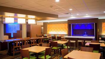 A hotel dining area with multiple tables and chairs, a bar area with a blue-lit backdrop, and a patterned wall.