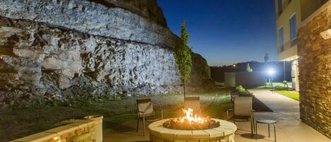 A stone fire pit with chairs and a table, set against a stone wall and a building in the background.