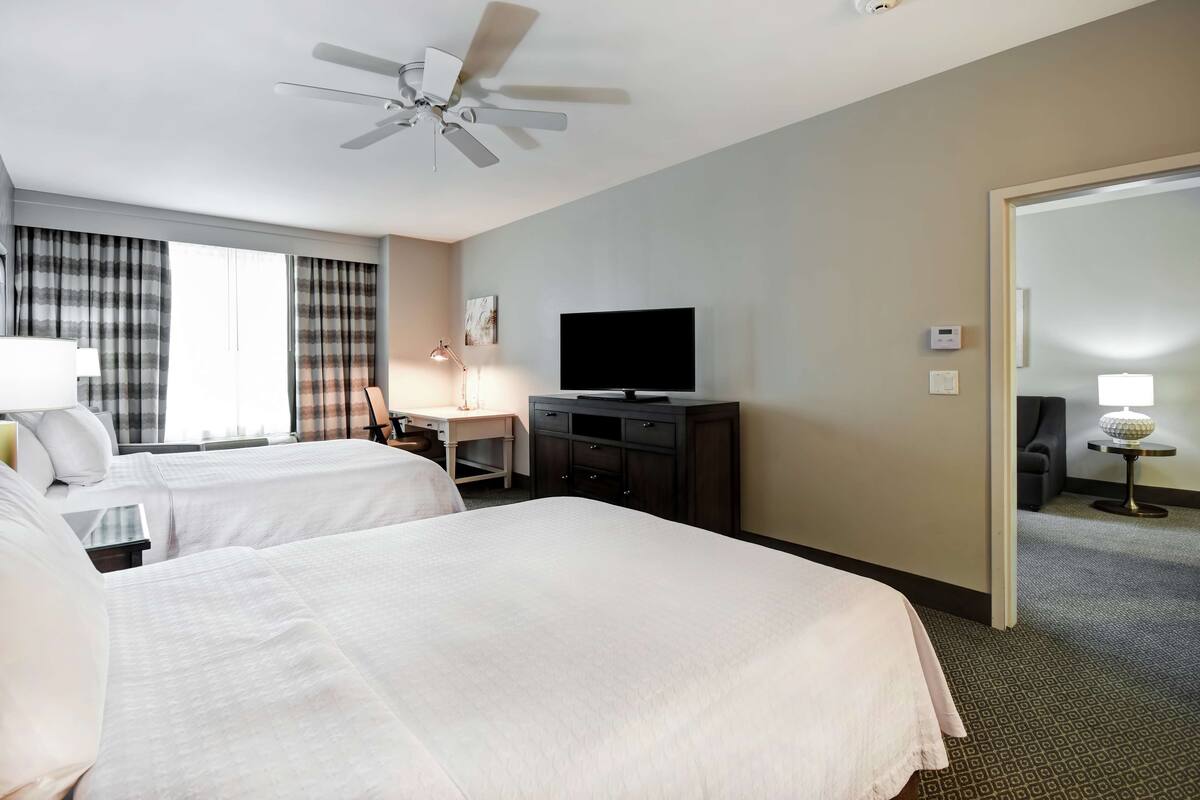 Une chambre d’hôtel avec deux lits, un bureau doté d’un téléviseur à écran plat, un ventilateur de plafond et une porte donnant sur une autre chambre.