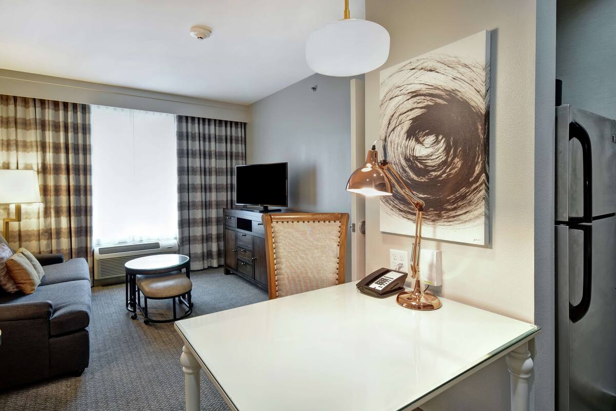 Une chambre d’hôtel équipée d’un canapé, d’une table à manger, d’un téléviseur, d’une lampe, d’un réfrigérateur et d’un grand tableau abstrait accroché au mur.