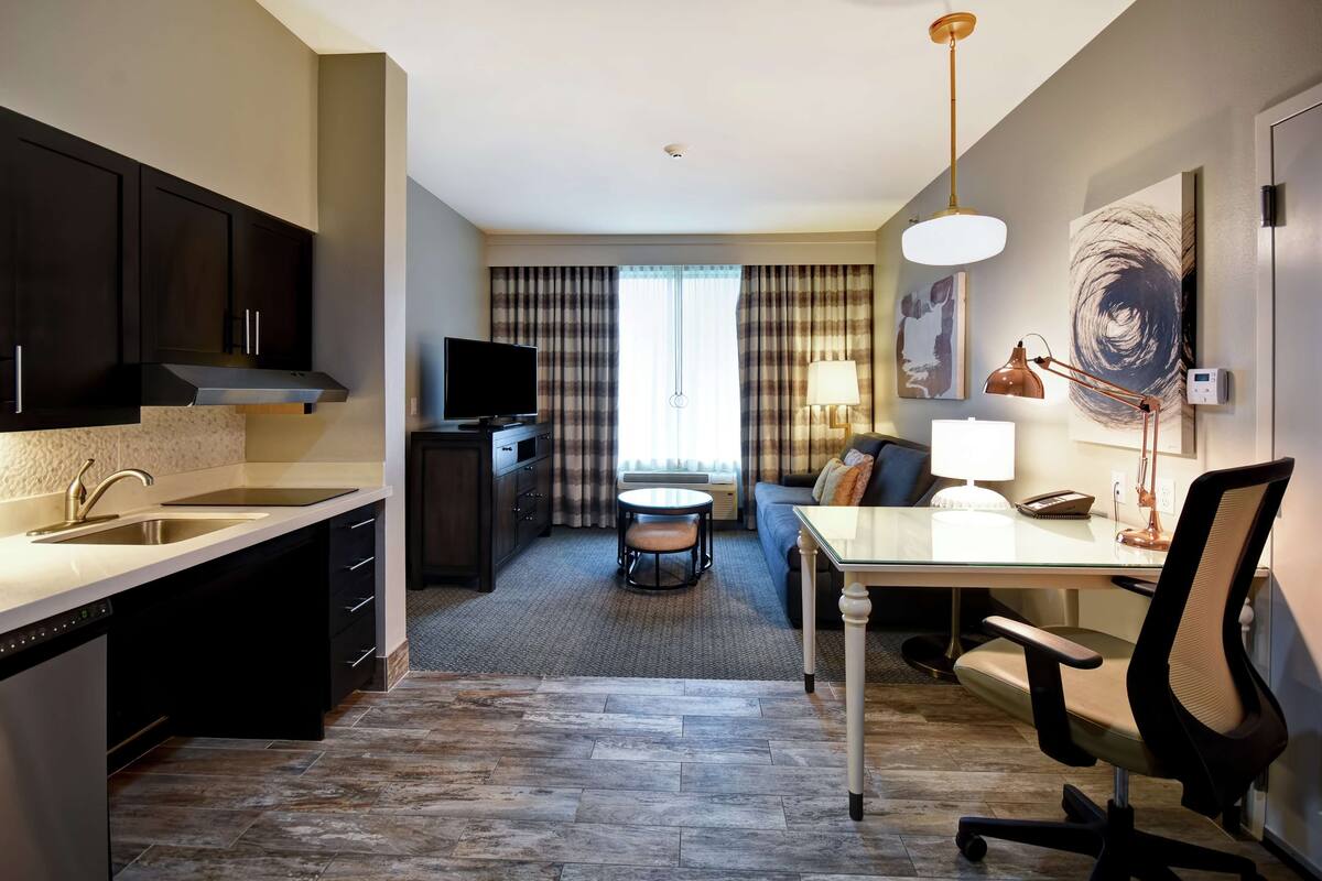 Une chambre d’hôtel moderne dotée d’une kitchenette, d’un coin repas, d’un canapé et d’une grande fenêtre avec des rideaux.