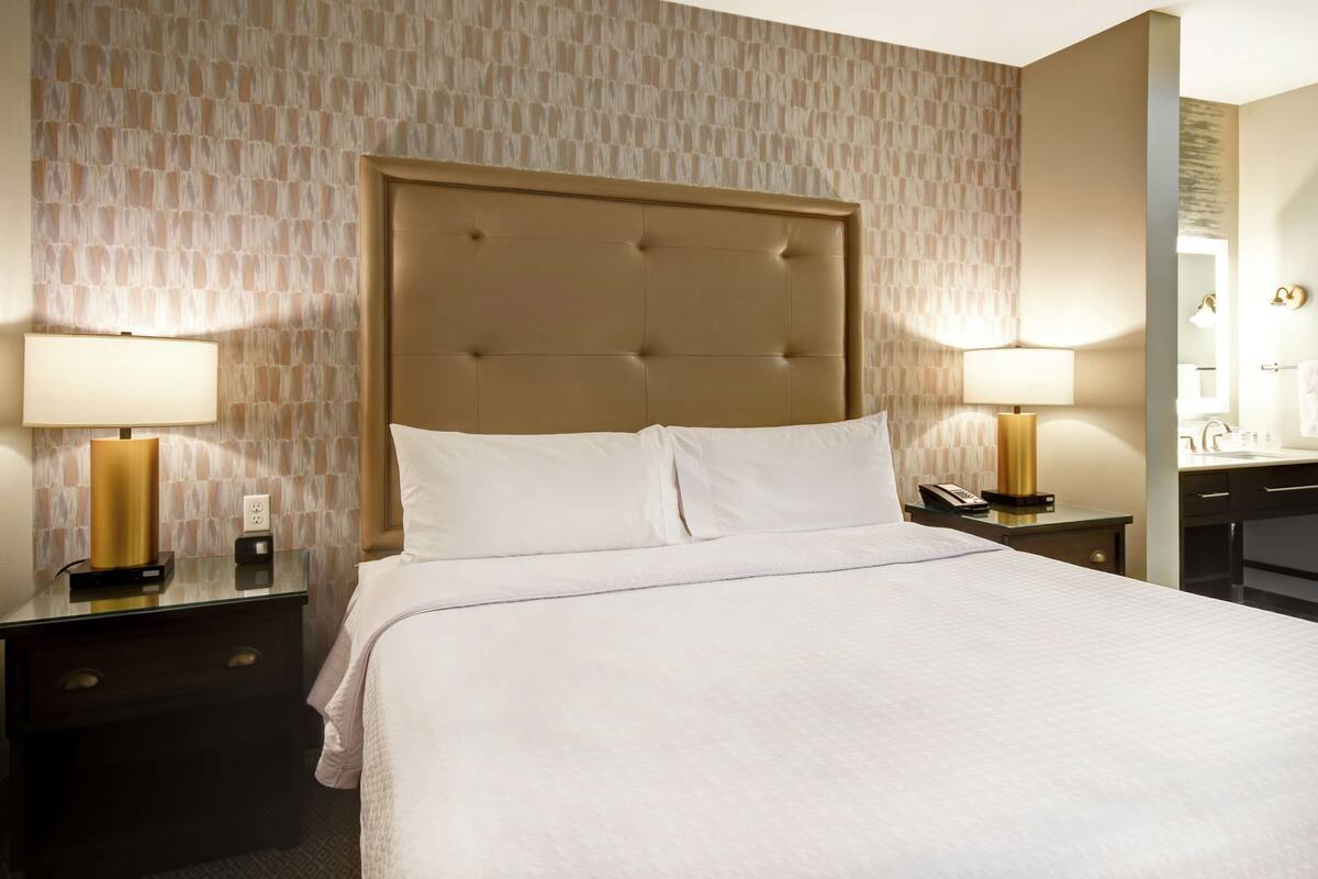 Une chambre d’hôtel avec un grand lit, deux tables de chevet avec des lampes, un miroir et une salle de bain visible à travers une porte ouverte.