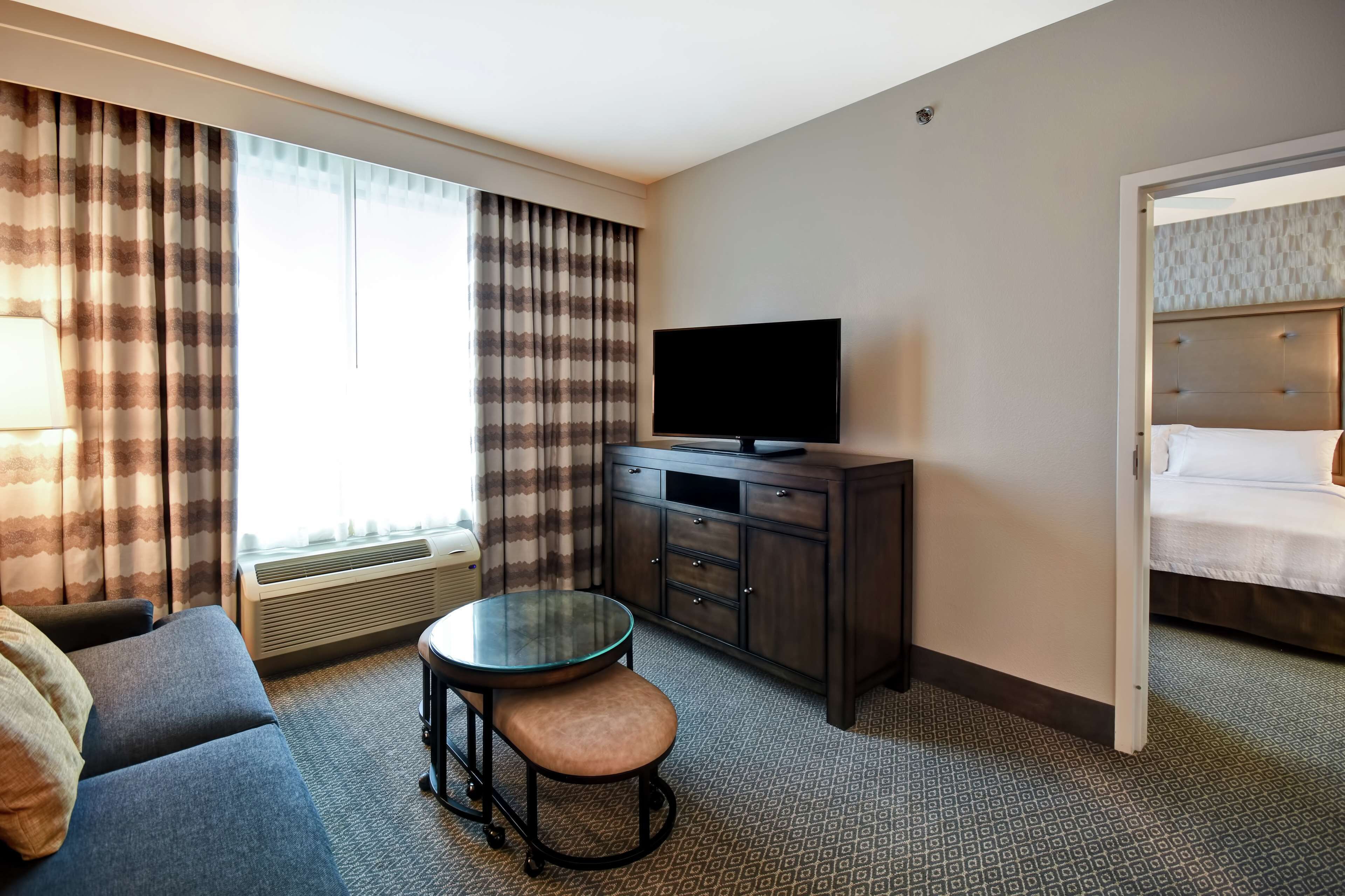 Ein Hotelzimmer mit einem Bett, einem Fernseher auf einer Holzkommode, einem Sofa, einem kleinen runden Tisch mit einem Hocker und einem Fenster mit Vorhängen.