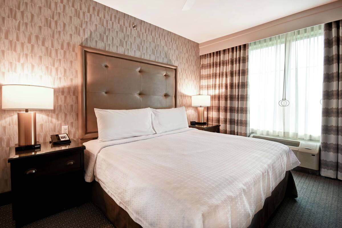 Une chambre d’hôtel avec un grand lit, deux tables de chevet avec des lampes, un téléphone fixé au mur et une fenêtre avec des rideaux.
