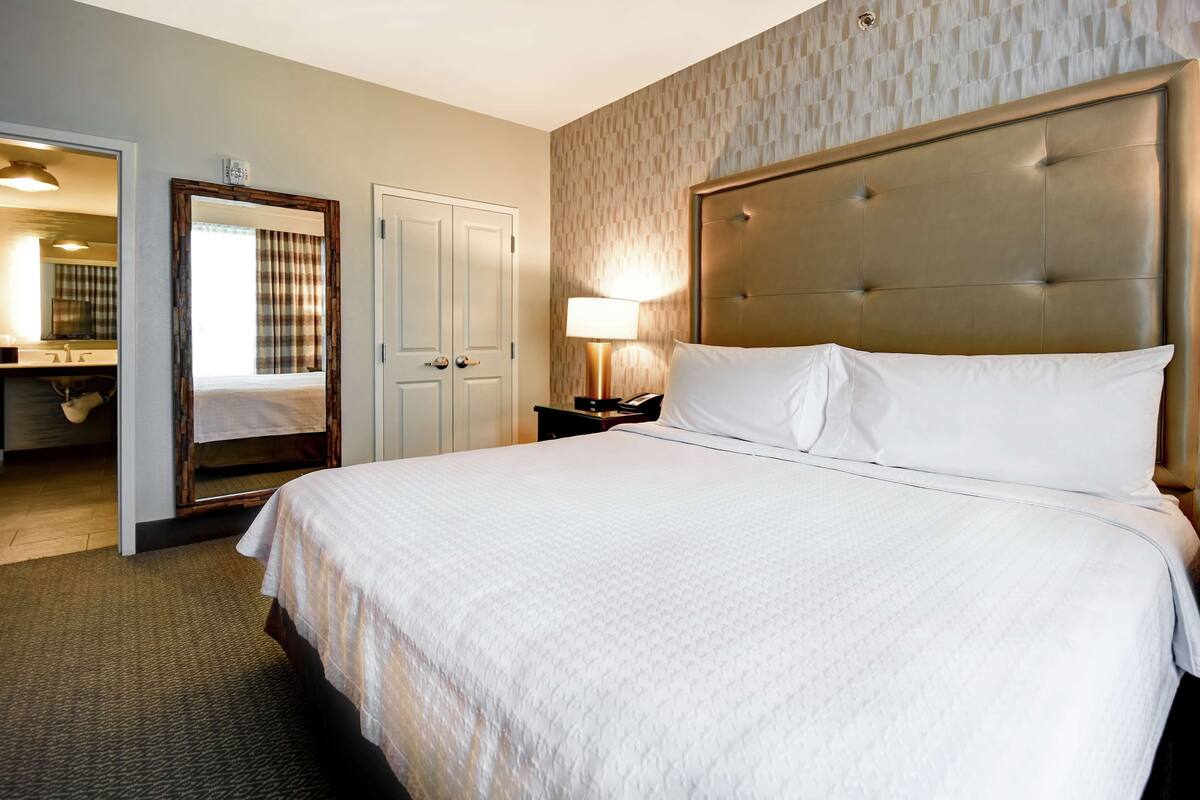 Une chambre d’hôtel avec un grand lit, une table de chevet avec une lampe, un miroir et une porte donnant sur une salle de bain.