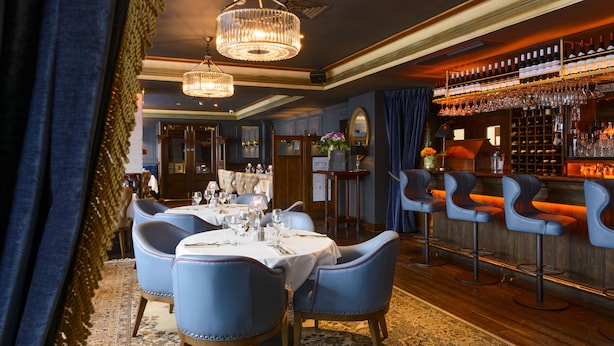 Een eethoek met een bar, met een kroonluchter, blauwe beklede stoelen en een wijnrek.