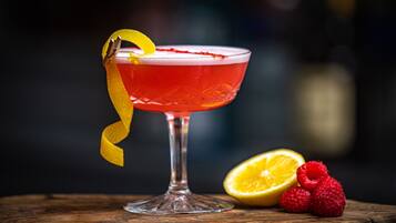 Een cocktail in een glas met een gele garnering, een halve citroen en frambozen op een houten ondergrond.