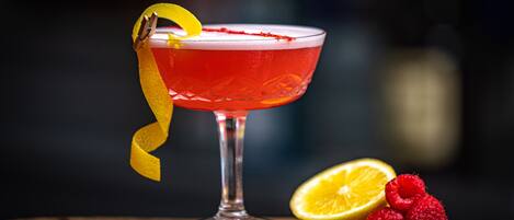 A cocktail in a glass with a yellow garnish, a half lemon, and raspberries on a wooden surface.