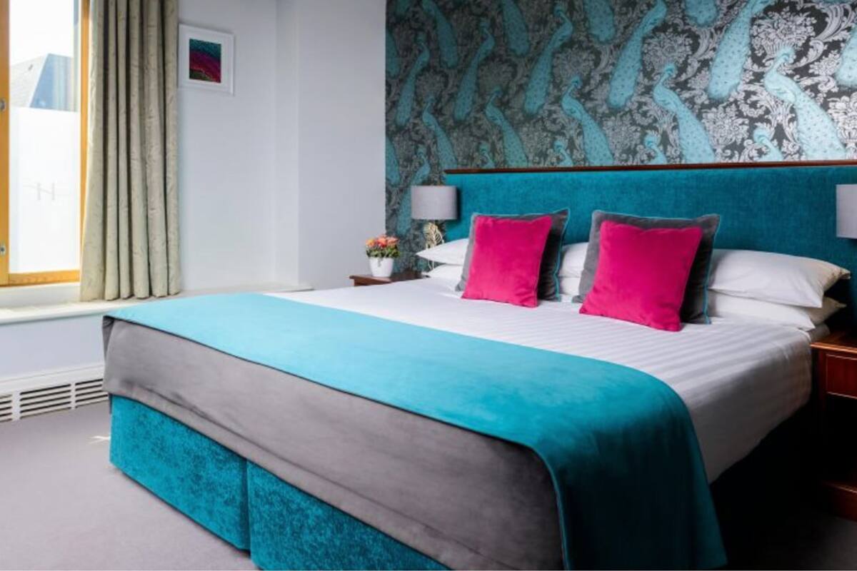 Ein Hotelzimmer mit einem großen Bett, blaugrünen und grauen Betttextilien, einem gemusterten Wandpapier und einem Fenster mit Vorhängen.