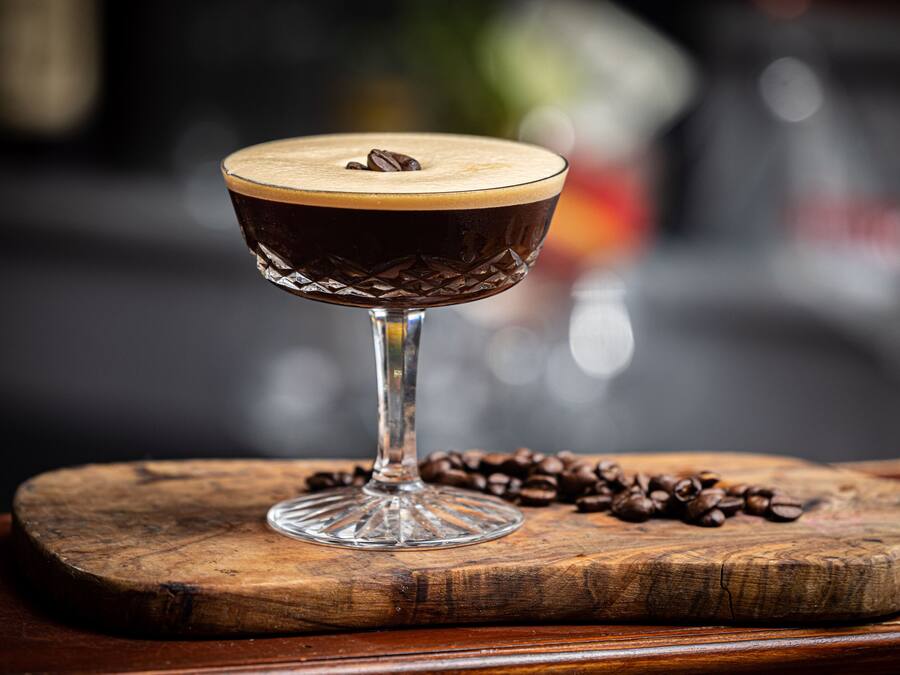 Un vaso con espuma en la parte superior, sobre un tablón de madera con granos de café esparcidos.