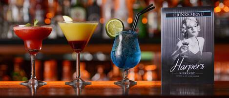 Tres cócteles sobre una barra con un menú de bebidas al fondo.