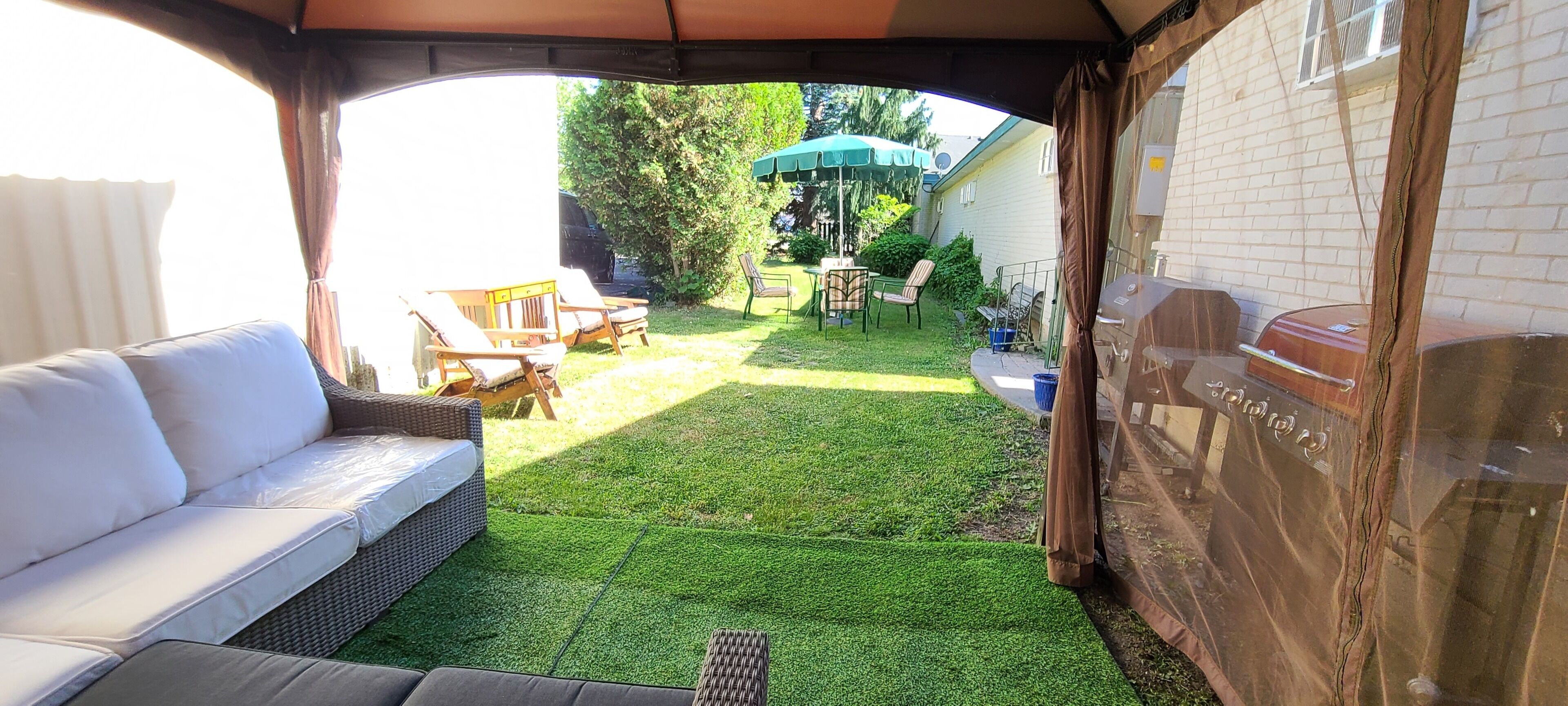 A covered outdoor seating area with a white sofa, a dining table with chairs, and a barbecue grill.