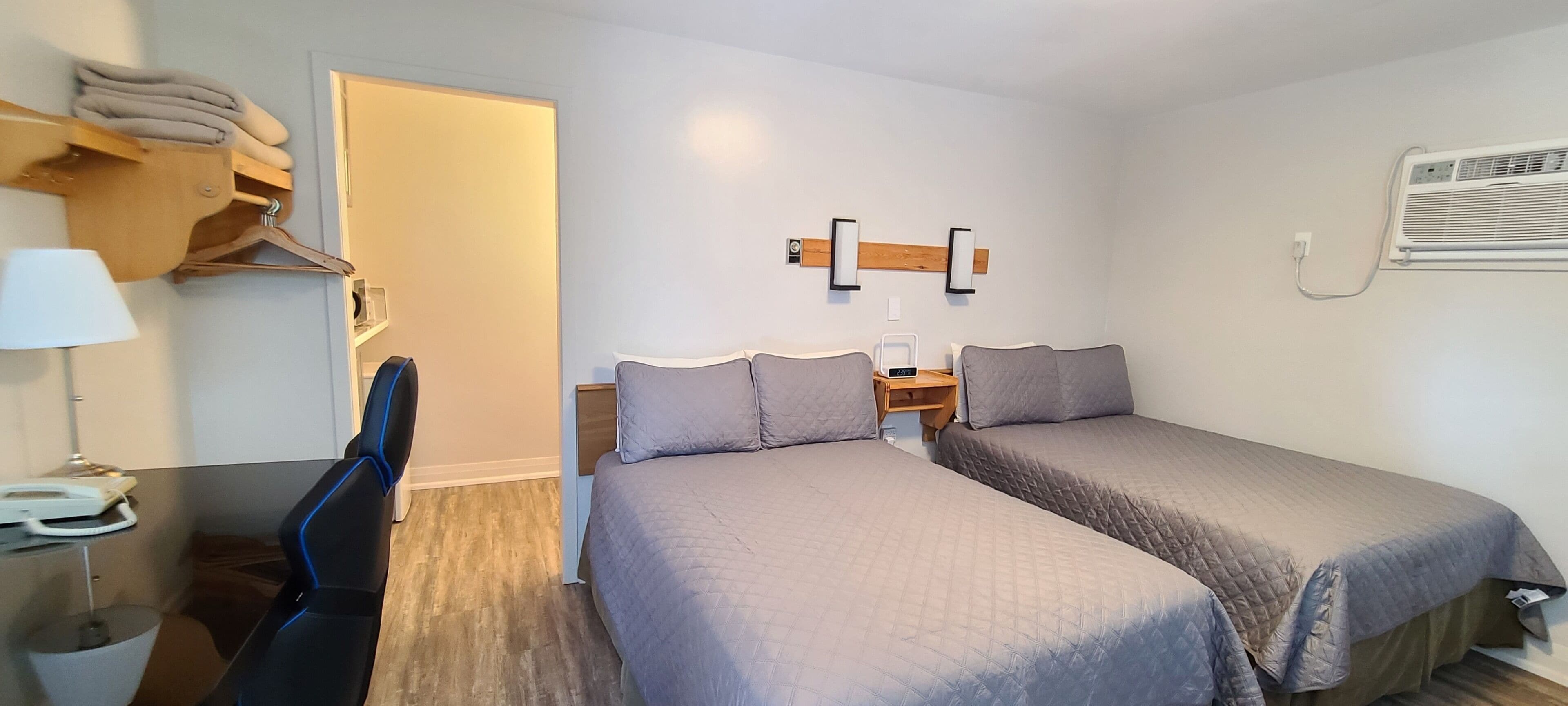 Ein Hotelzimmer mit zwei Betten, einem Schreibtisch, einem Stuhl, einer Lampe, einem Kleiderschrank und einer Klimaanlage.