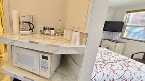 A compact kitchenette with a microwave, coffee maker, and a small fridge. There is a TV and a window with blinds in the background. - Maple Leaf Motel (Goderich)