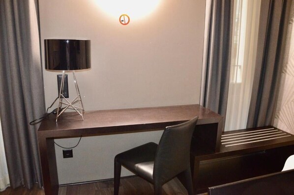 A hotel room with a desk, a chair, a lamp, and a window with curtains.