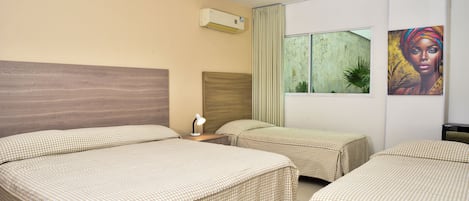 A hotel room with three beds, a window with a view, an air conditioning unit, and a painting on the wall.