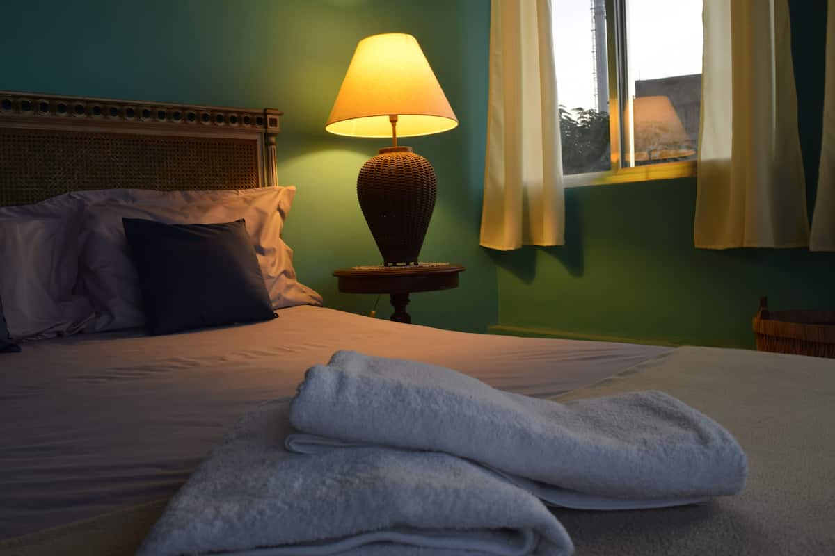 A neatly made bed with a folded towel, a lamp on a bedside table, and a window with curtains.