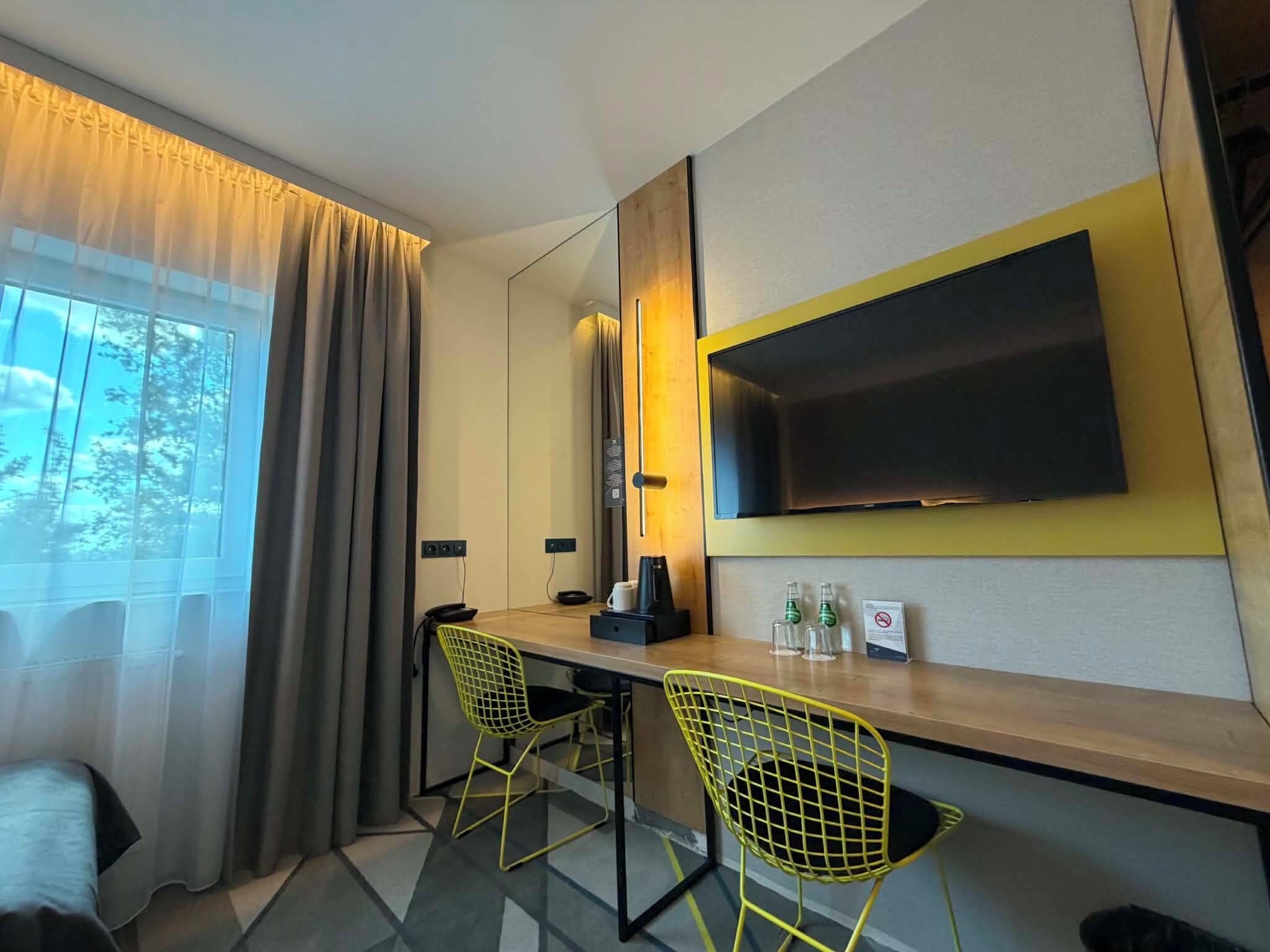 Habitación de hotel con un ventanal grande, un televisor de pantalla plana montado en la pared, un escritorio con dos sillas y una lámpara.