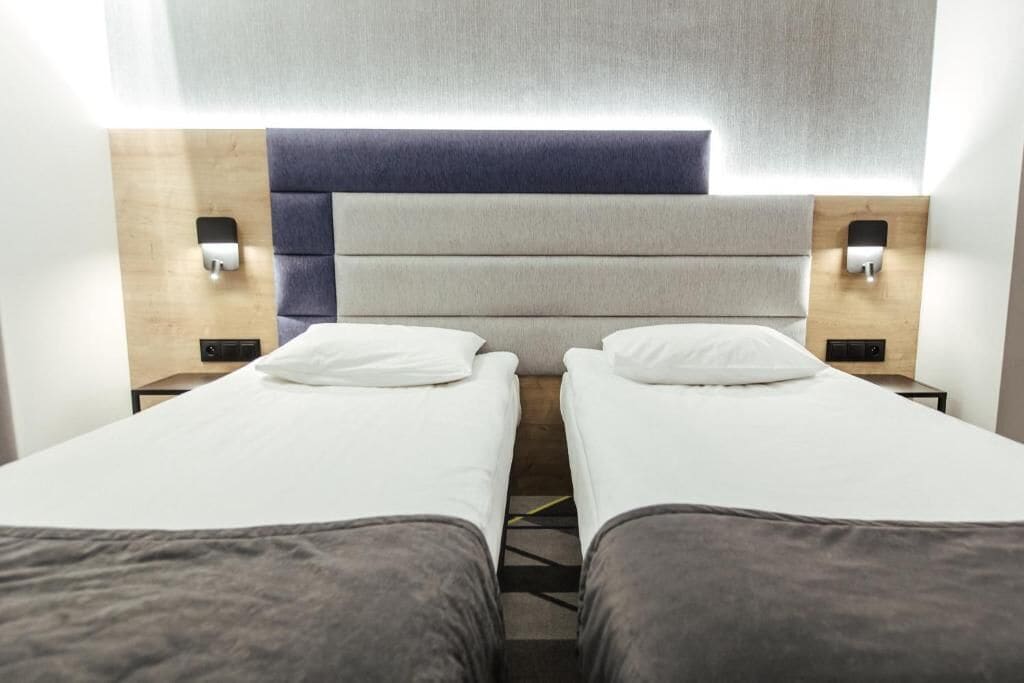 A hotel room with two single beds, a wooden headboard, bedside lamps, and a neutral color scheme.
