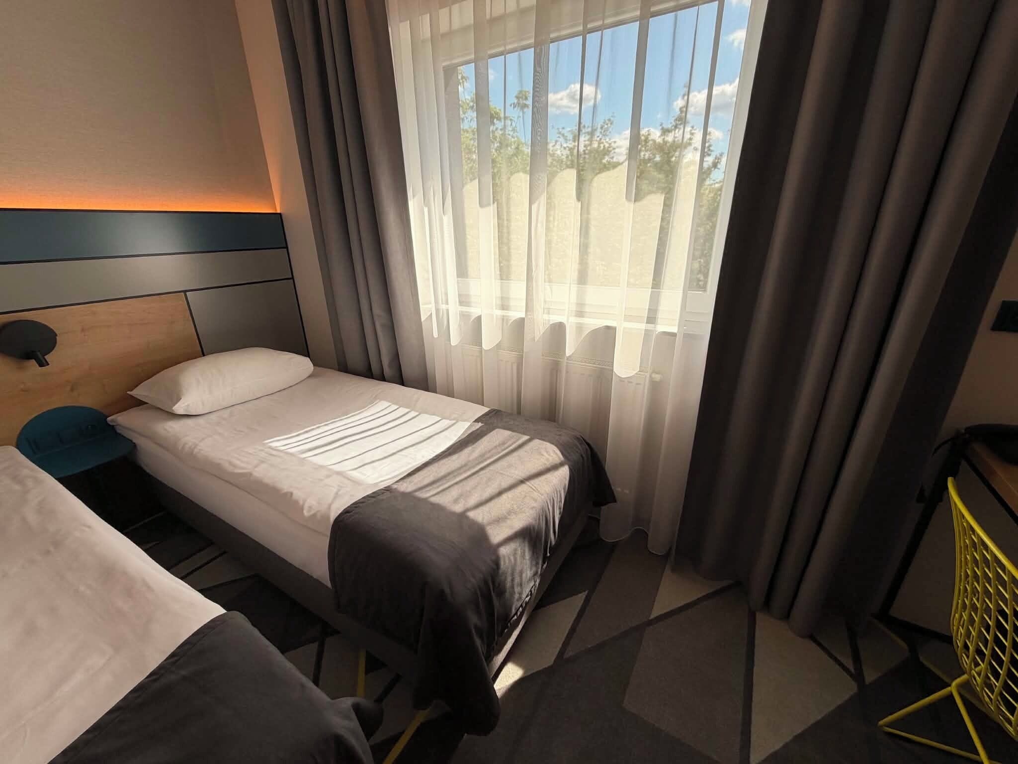 Hotellihuone, jossa on yksi sänky, yöpöytä, tuoli ja ikkuna, jossa on verhot.