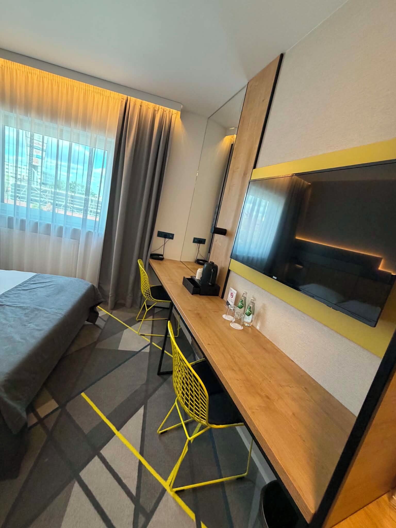 Habitació d'hotel amb llit, escriptori, cadira, televisió i finestra amb cortines.