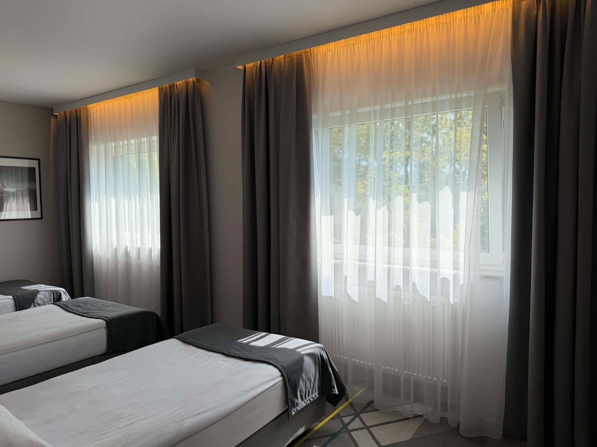 A hotel room with two beds, a large window with sheer curtains, and a framed picture on the wall.