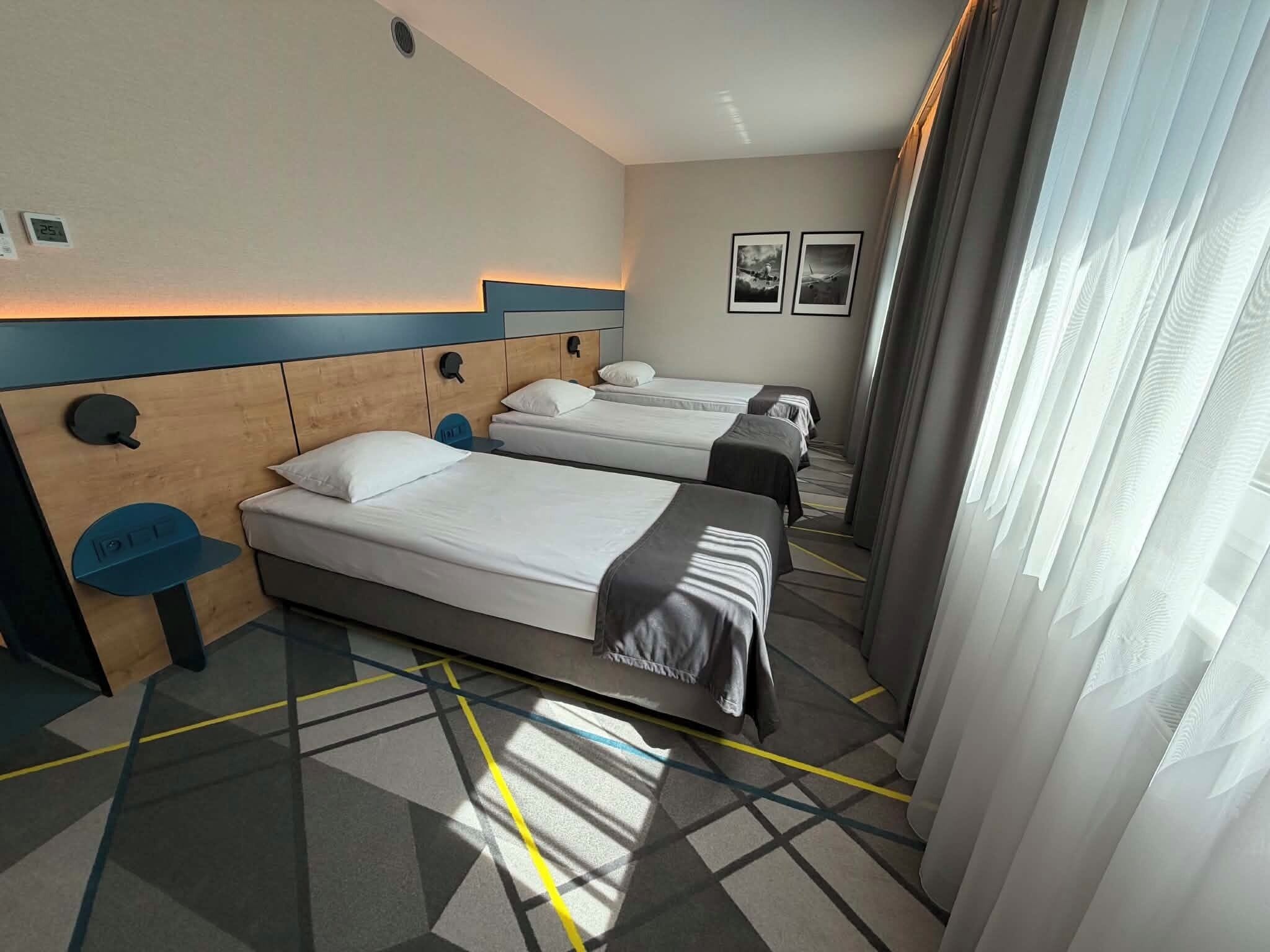 A hotel room with two beds, a headboard with a light fixture, a small table, and a window with curtains.
