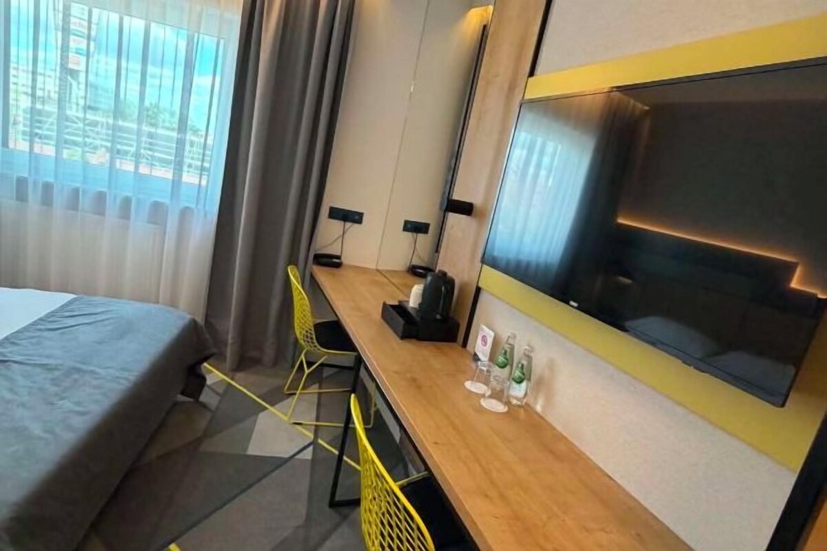 Habitación de hotel con cama, escritorio, silla, televisor y ventana con cortinas.