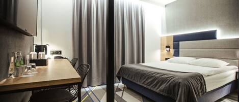 A hotel room with a bed, a desk with chairs, a TV, and a window with curtains.