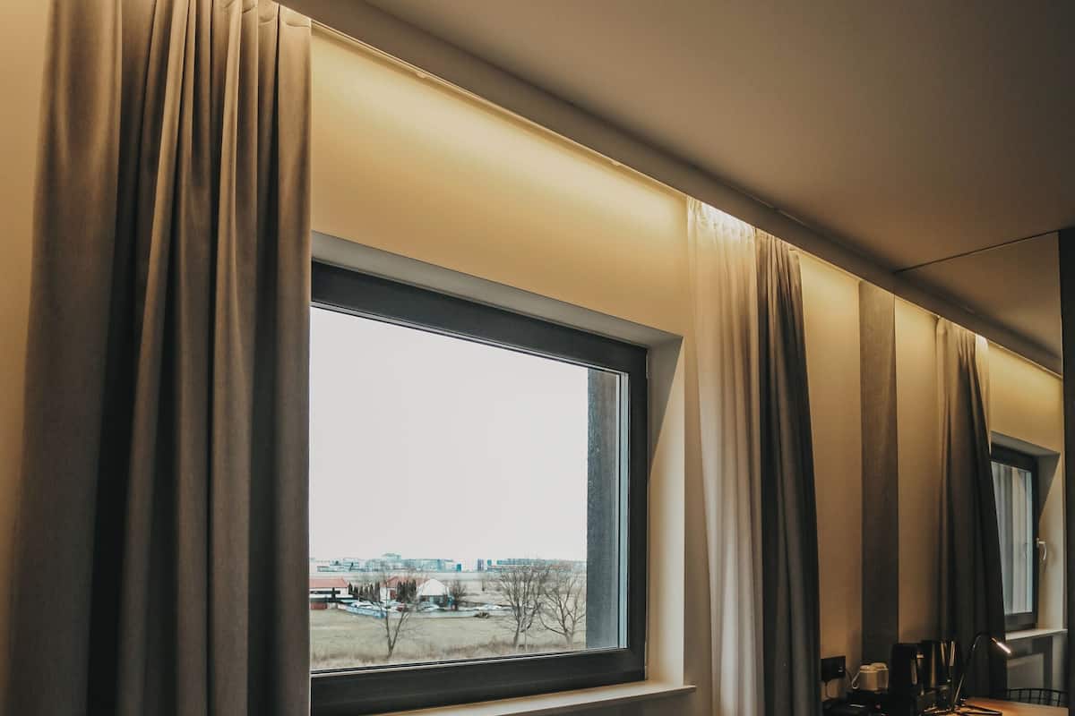 A hotel room with a bed, a desk with a chair, a window with curtains, and a radiator.