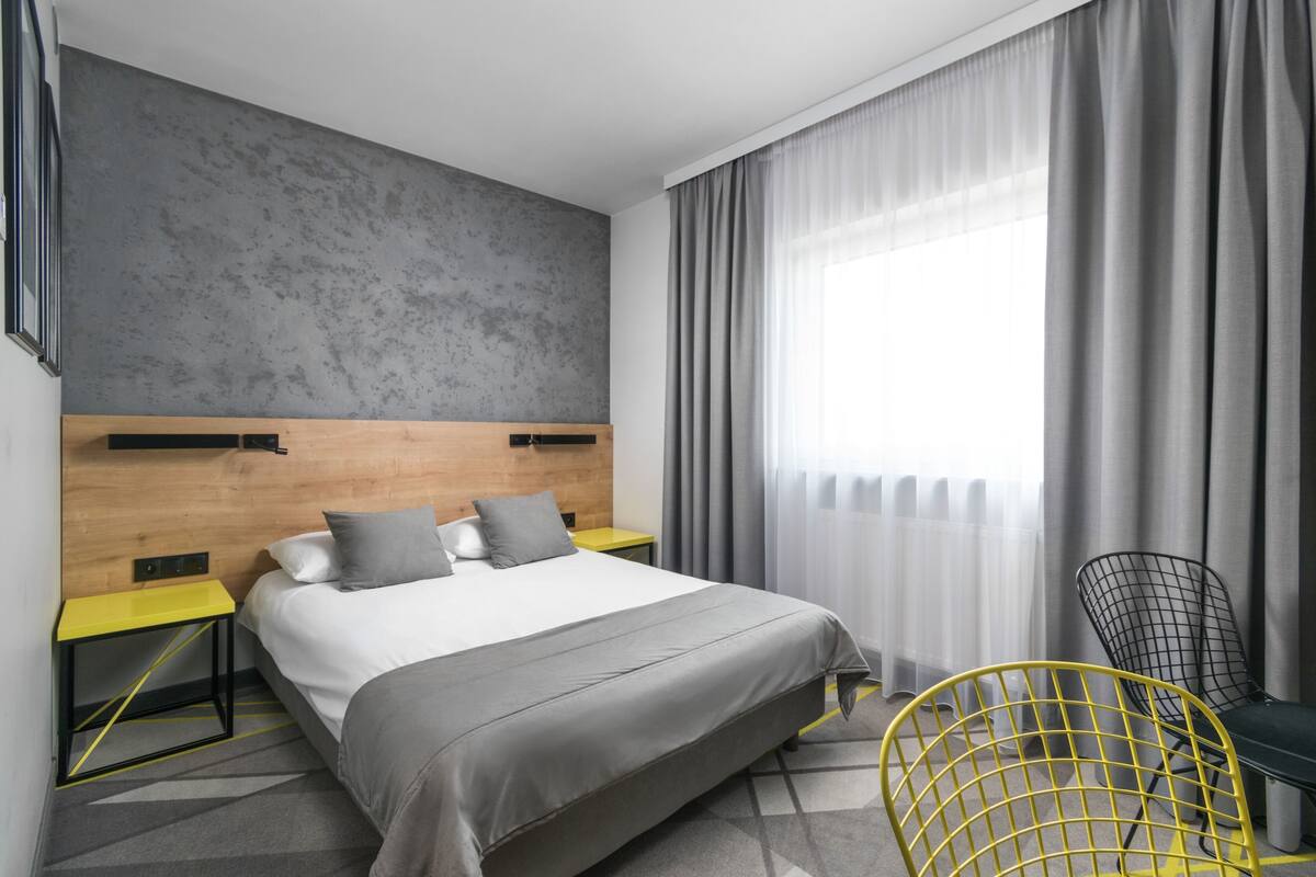 A modern hotel room with a large bed, a wooden headboard, a yellow chair, and a view of the outdoors through sheer curtains.