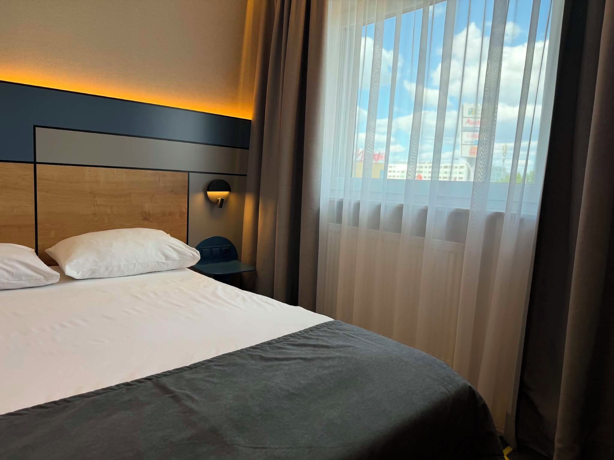 A hotel room with a bed, a nightstand, and a window with curtains.