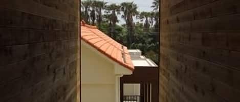 A wooden interior hallway leading to a balcony with a red roof, a small outdoor area with steps, and a view of palm trees.