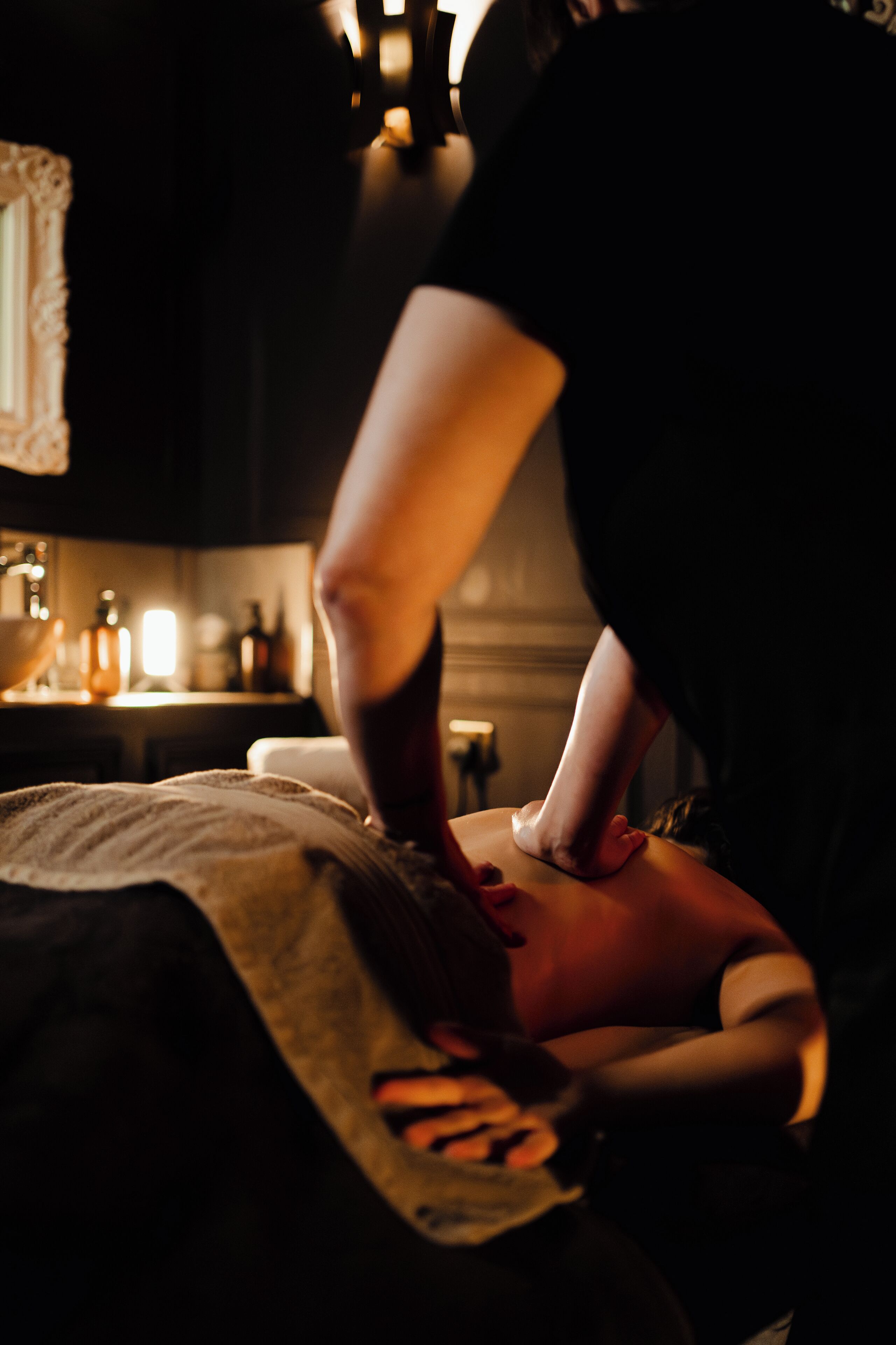 A massage therapy session in a dimly lit room with a wooden massage table, a towel, and a bottle of oil on the table.