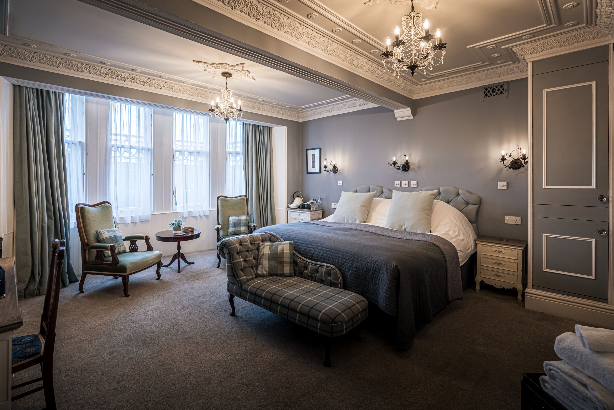 A spacious bedroom with a large bed, a chaise lounge, a small table, and a chandelier.