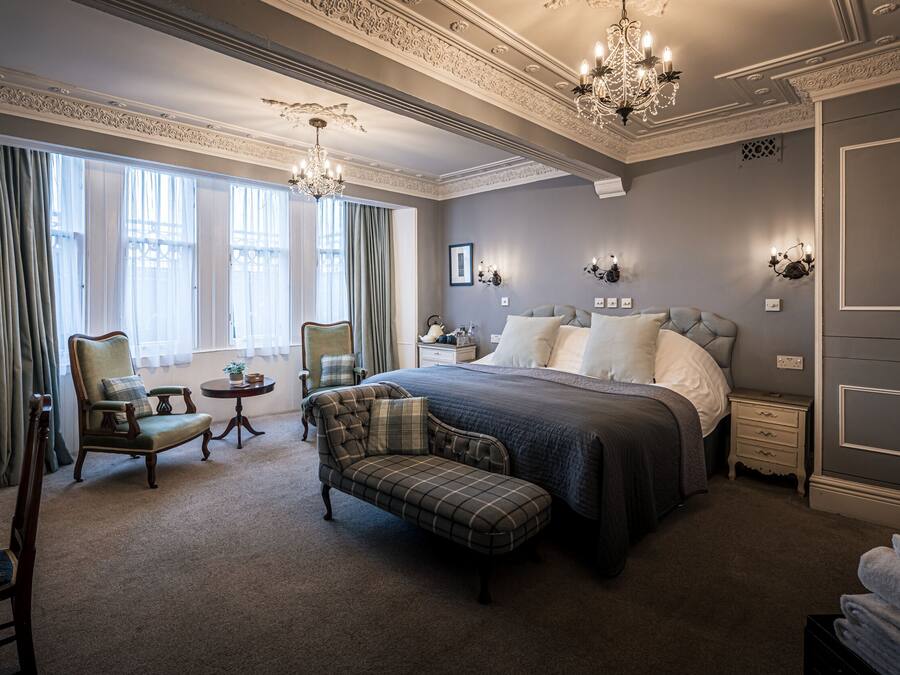 A spacious bedroom with a large bed, a chaise lounge, a small table, and a chandelier.